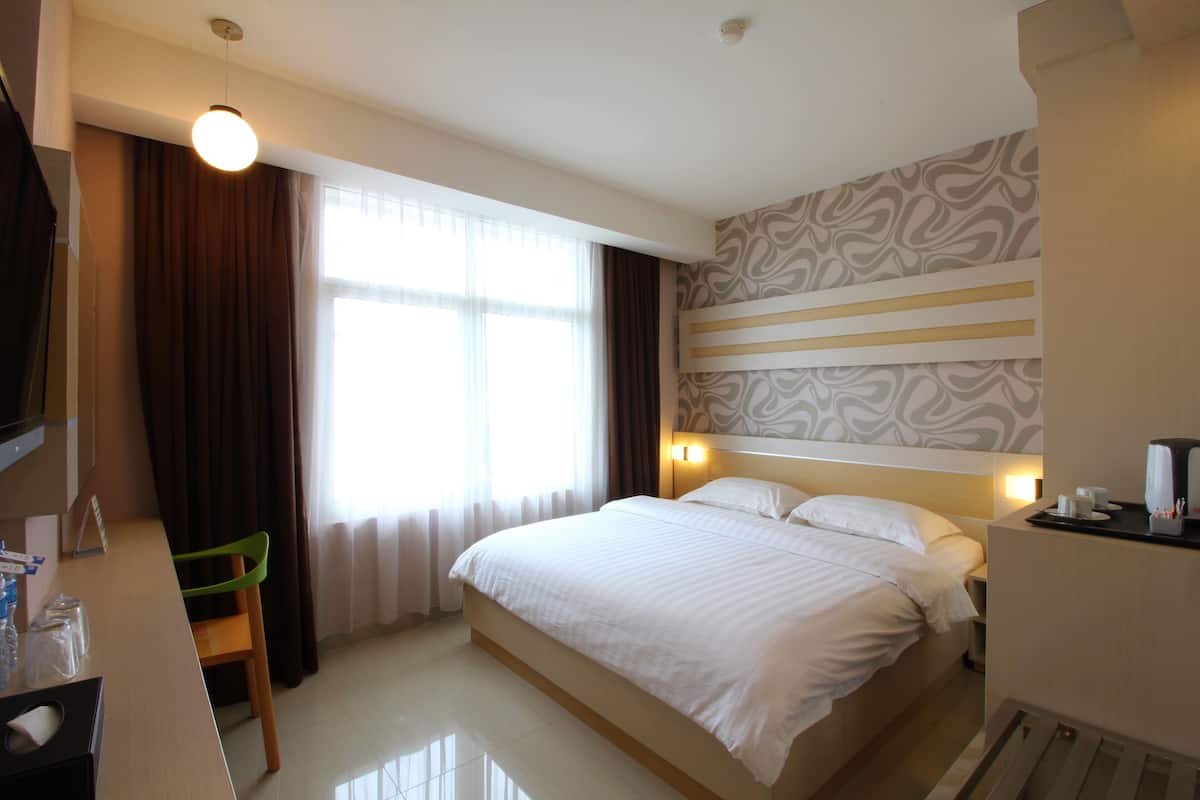 A hotel room with a bed, a desk, a chair, a window with curtains, and a patterned wall.