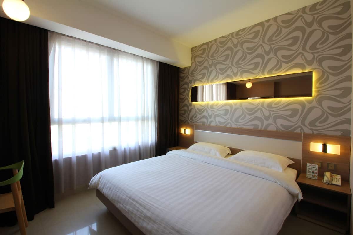 A hotel room with a large bed, a bedside table with a lamp, a wall-mounted mirror, and a window with curtains.