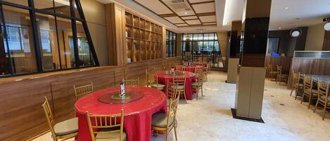 A spacious restaurant with round tables and chairs, wooden paneling, and large windows.
