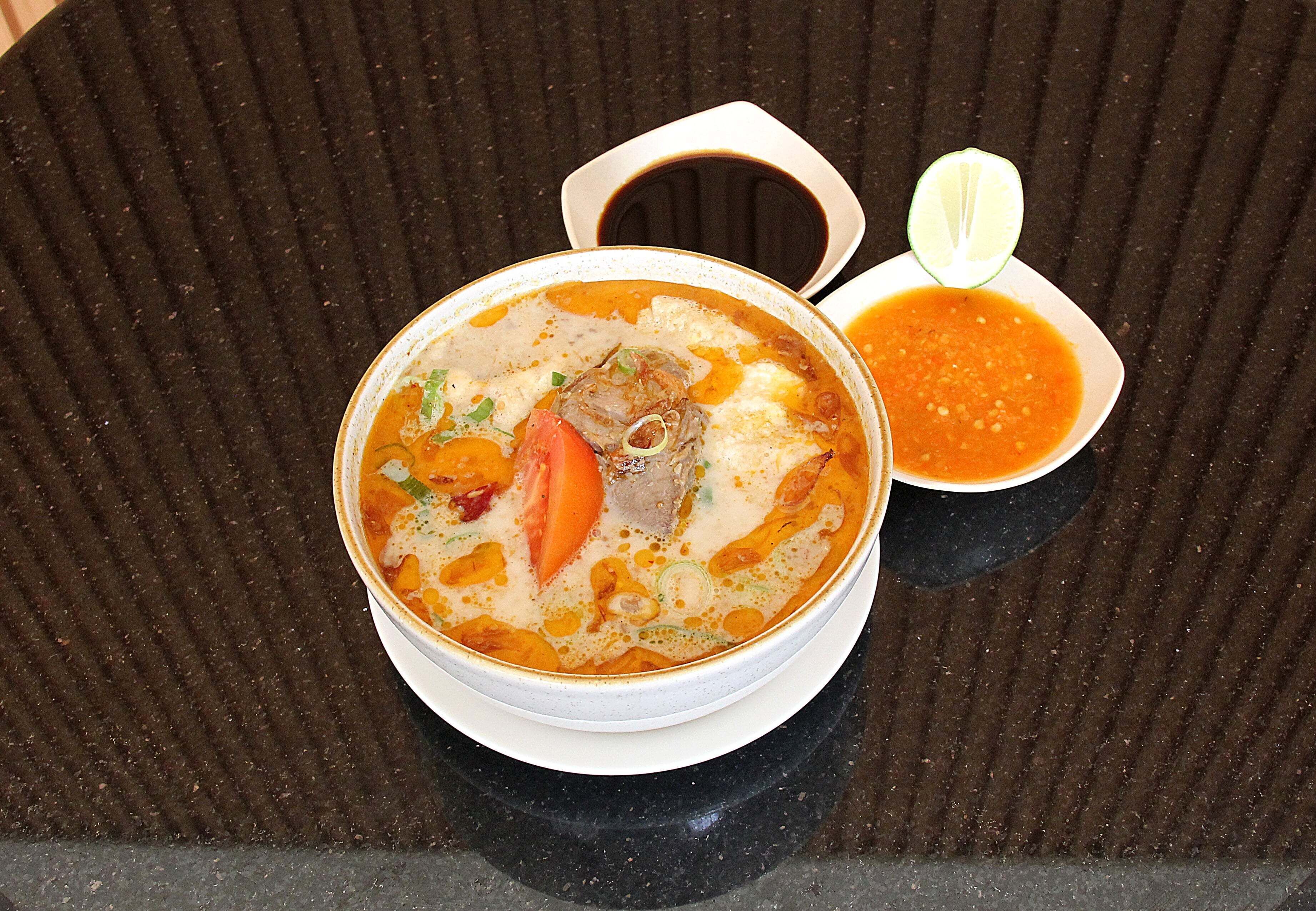 A bowl of soup with a side of sauce and a lime wedge on a textured black surface.