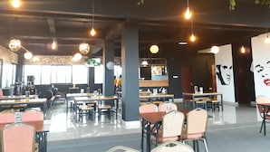 A spacious cafe with multiple tables and chairs, hanging pendant lights, and a mural on the wall. - Urban Style Hotel Lampung, Azana Hotels Collection (Gedongtataan)