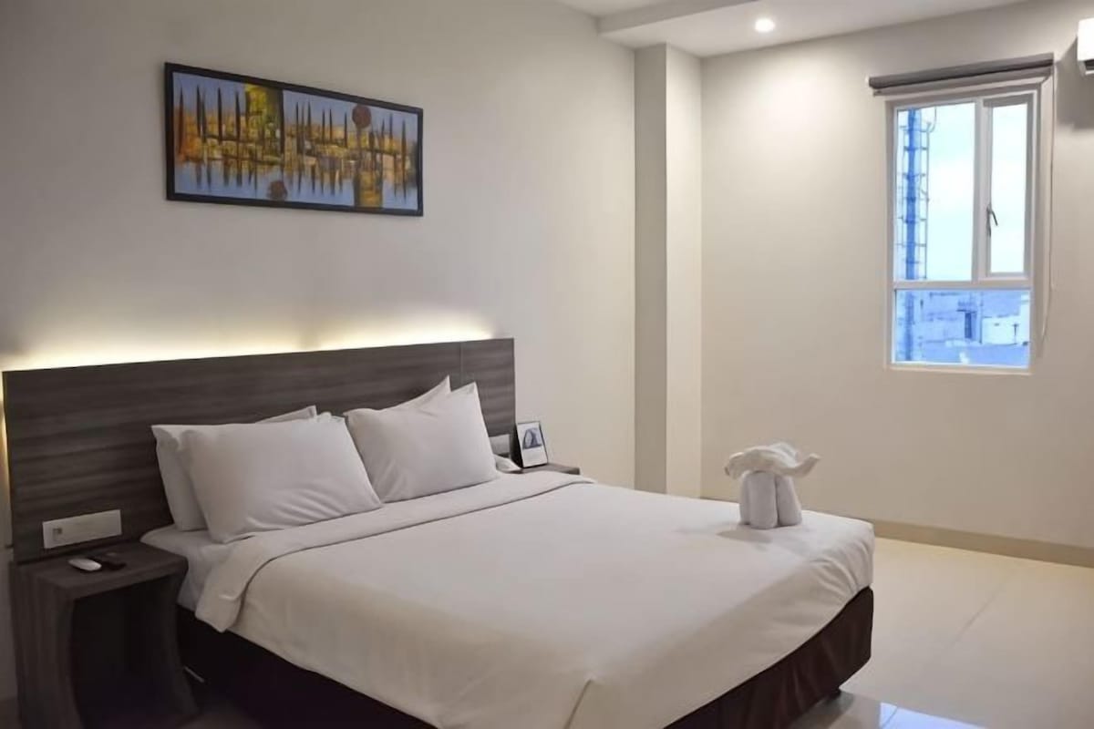 A neatly made bed with white linens, a bedside table with a lamp, a framed picture on the wall, and a window with blinds.