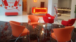 A lobby with orange chairs, a reception desk, and a wall with a logo. - Urban Style Hotel Lampung, Azana Hotels Collection (Gedongtataan)