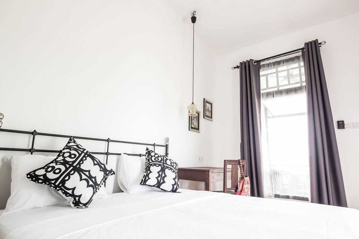 Un lit avec des coussins à motifs, une table de chevet, une chaise, une lampe suspendue et une fenêtre avec des rideaux.