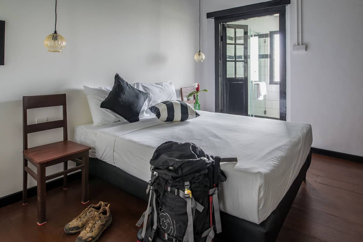 Un lit bien fait, avec du linge de lit blanc, une chaise en bois, un sac à dos et une paire de chaussures posées sur le sol.