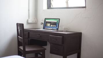 A small, dark wooden desk with a laptop, a white mug, and a chair in a room with white walls and a window.
