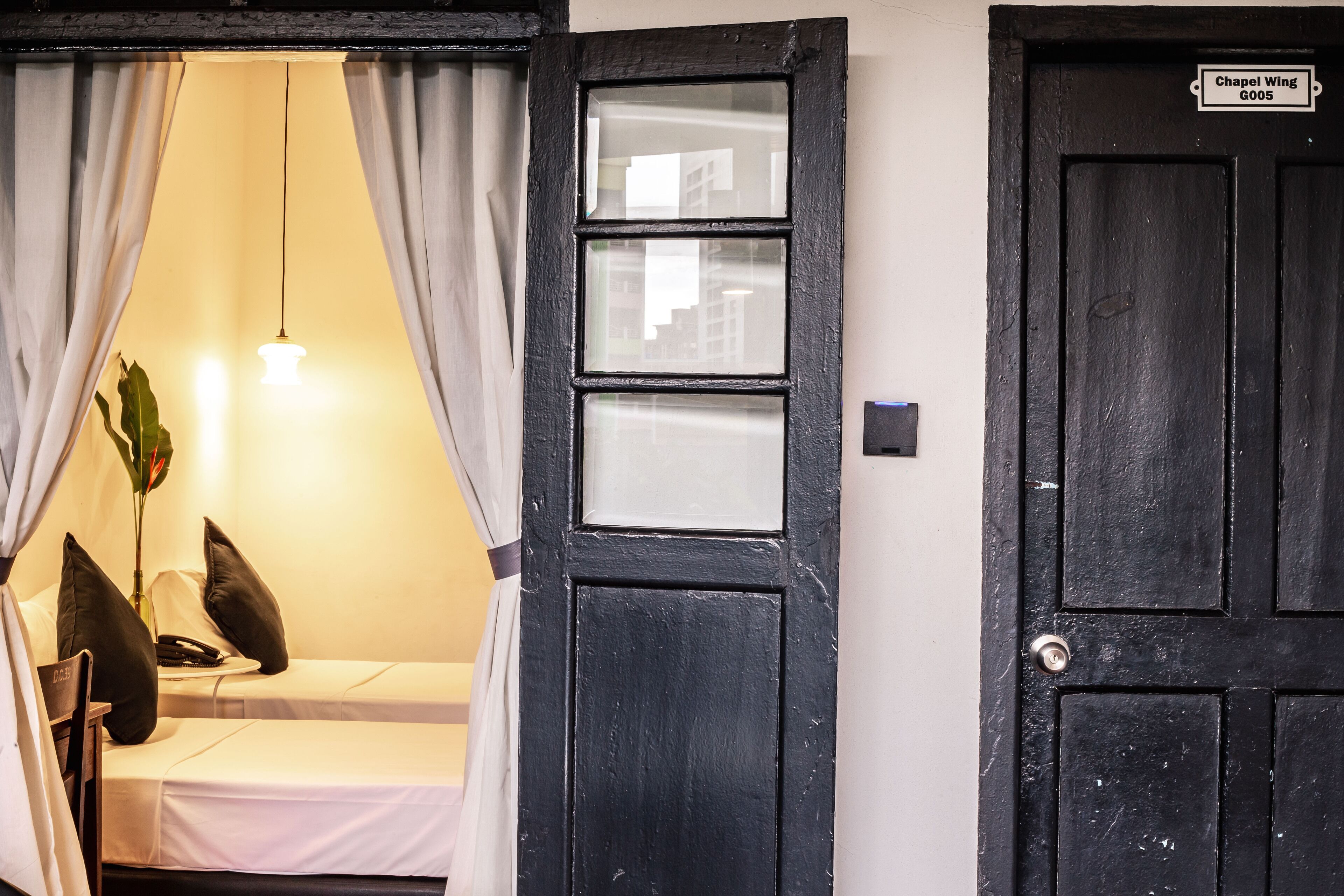 A room with a bed, a bedside table with a lamp, and a door labeled "Chapel Wing G005".