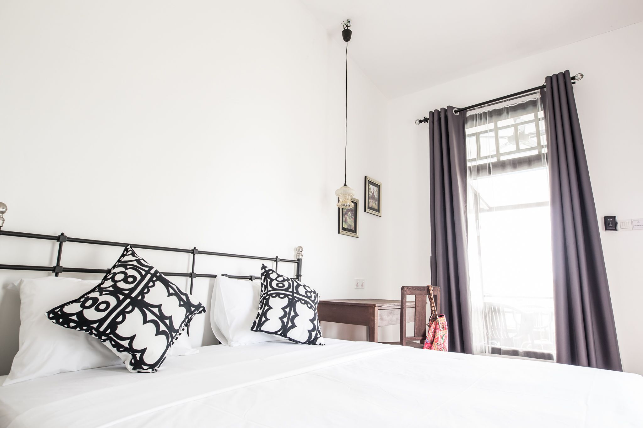 A neatly made bed with patterned pillows, a hanging light fixture, and a view of the outdoors through a window with curtains.