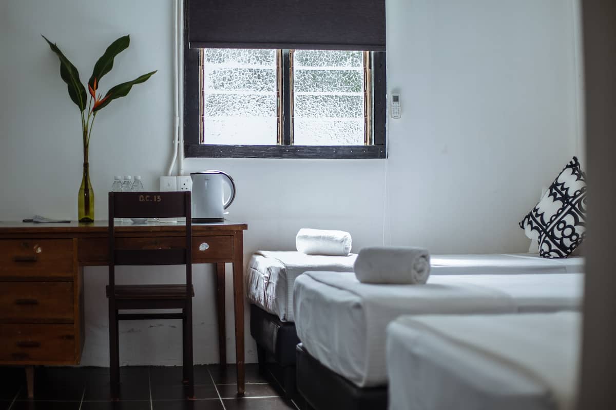 Une chambre d’hôtel équipée d’un bureau, d’une chaise, de deux lits, d’une fenêtre, d’une plante et d’un coussin décoratif.