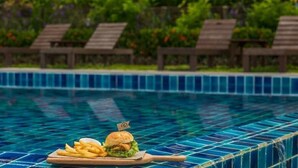 Zona de comedor junto a la piscina con una mesa preparada para una comida, incluyendo una hamburguesa y papas fritas, y sillones de descanso al fondo.