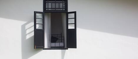 A room with a single bed, a window with open shutters, and a white wall.