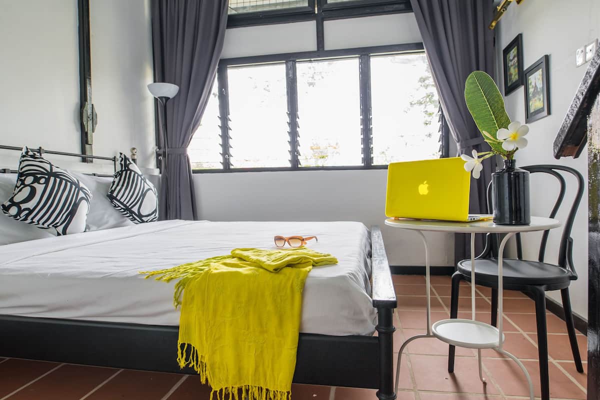 Une chambre à coucher avec un lit, un ordinateur portable posé sur une petite table, une chaise et une fenêtre avec des rideaux.