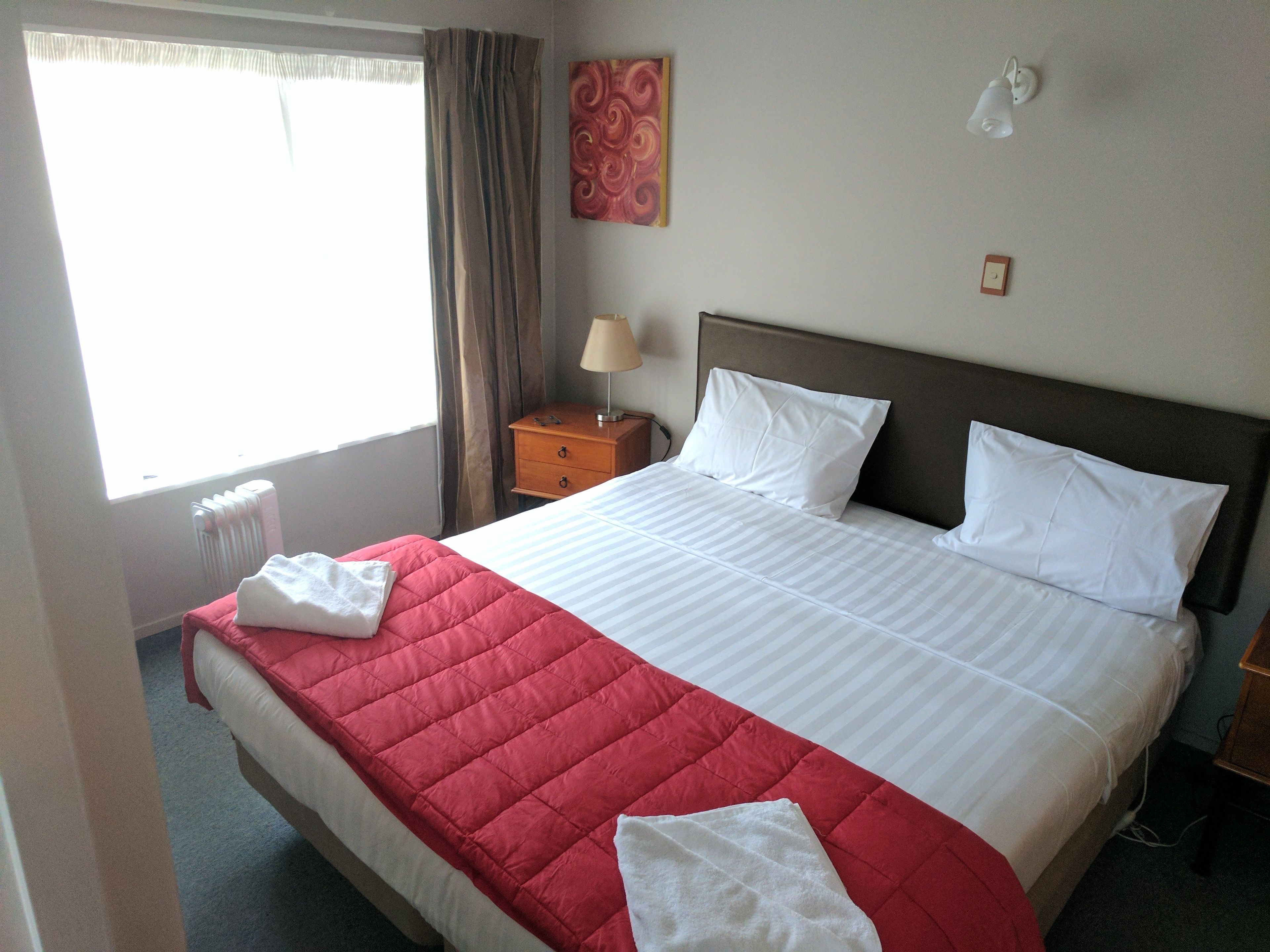Habitación de hotel con cama, mesita de noche, lámpara, radiador y ventana con cortinas.