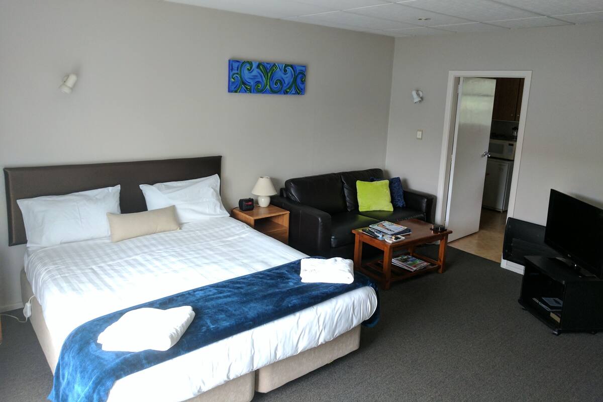A hotel room with a bed, a sofa, a TV, and a door leading to a kitchen.
