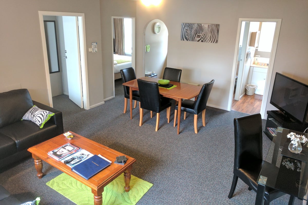 A living room with a sofa, coffee table, dining table with chairs, a TV, and a kitchen in the background.