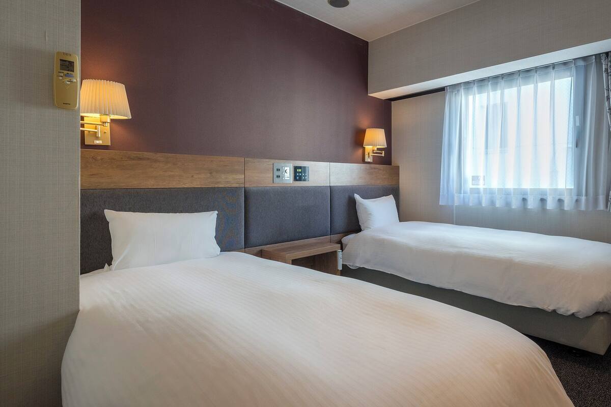 A hotel room with two single beds, a wooden headboard, a control panel, and a window with sheer curtains.