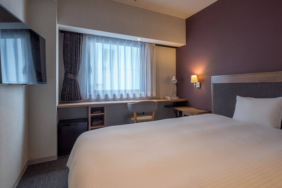 A hotel room with a bed, a desk, a chair, a TV, and a window with curtains.