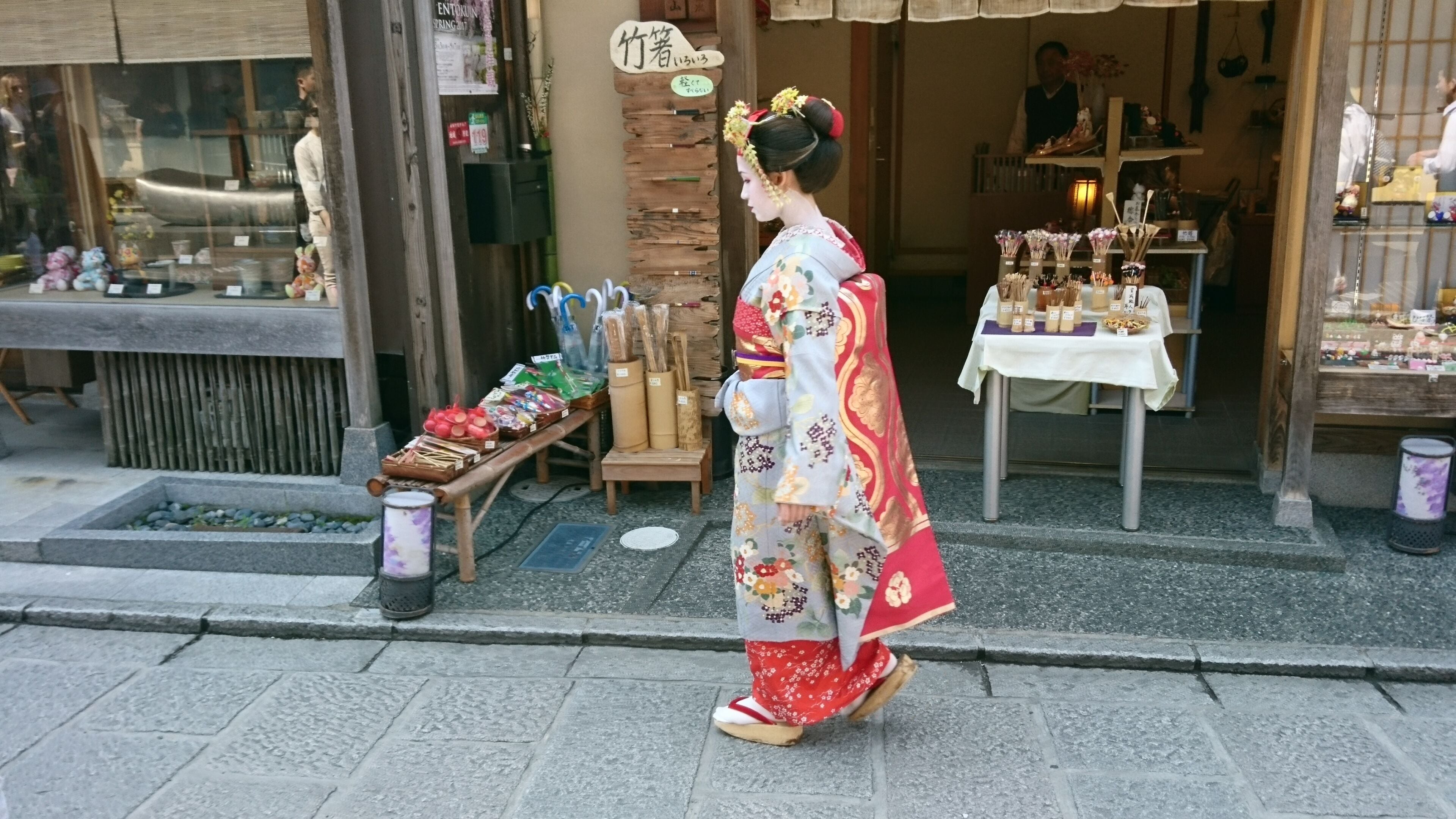 A traditional Japanese street scene with a person in a kimono walking past a shop displaying various goods.