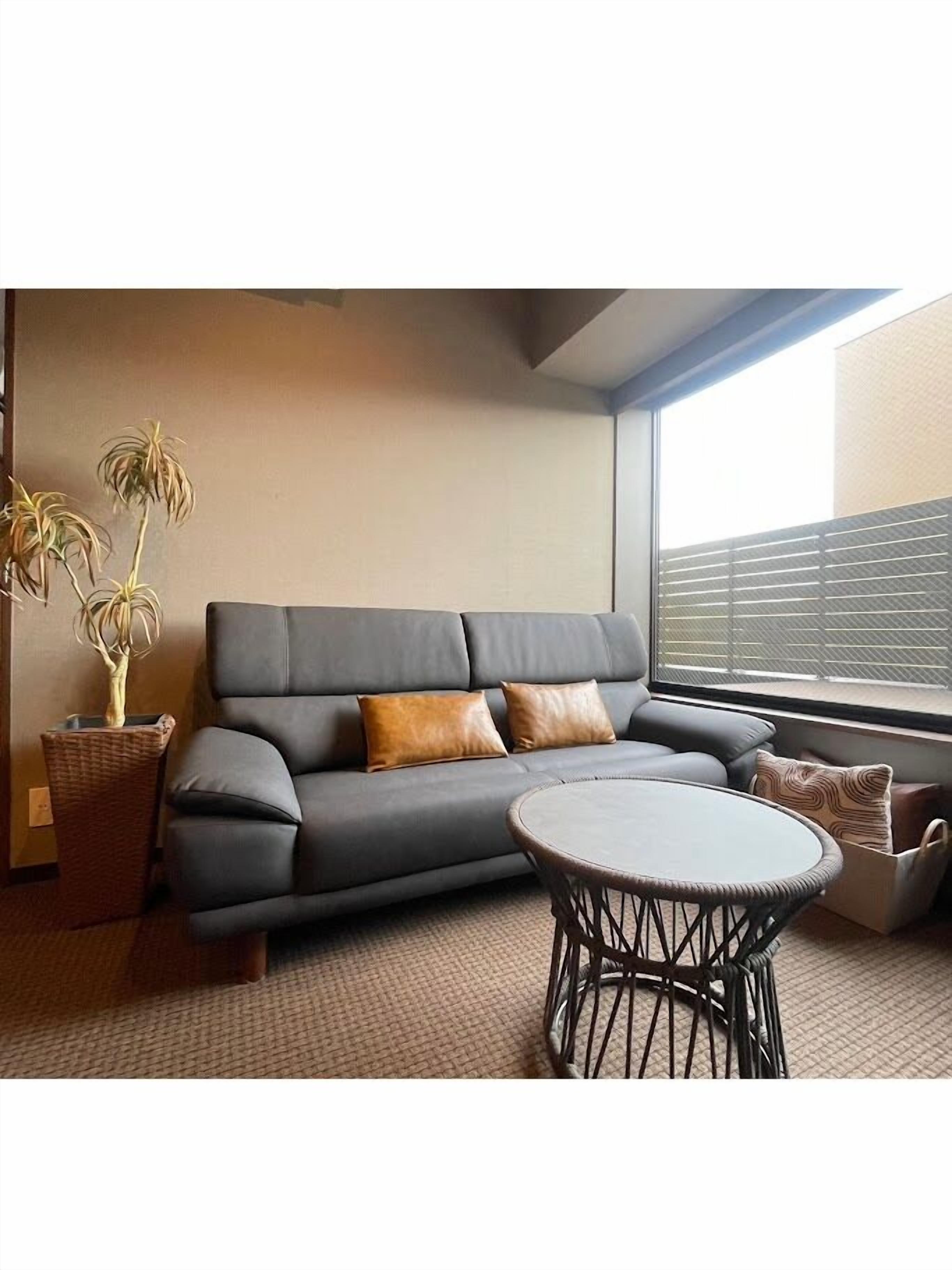 Ein modernes Wohnzimmer mit einem grauen Sofa, zwei braunen Kissen, einem runden Korbtisch und einem Topf mit Pflanze.