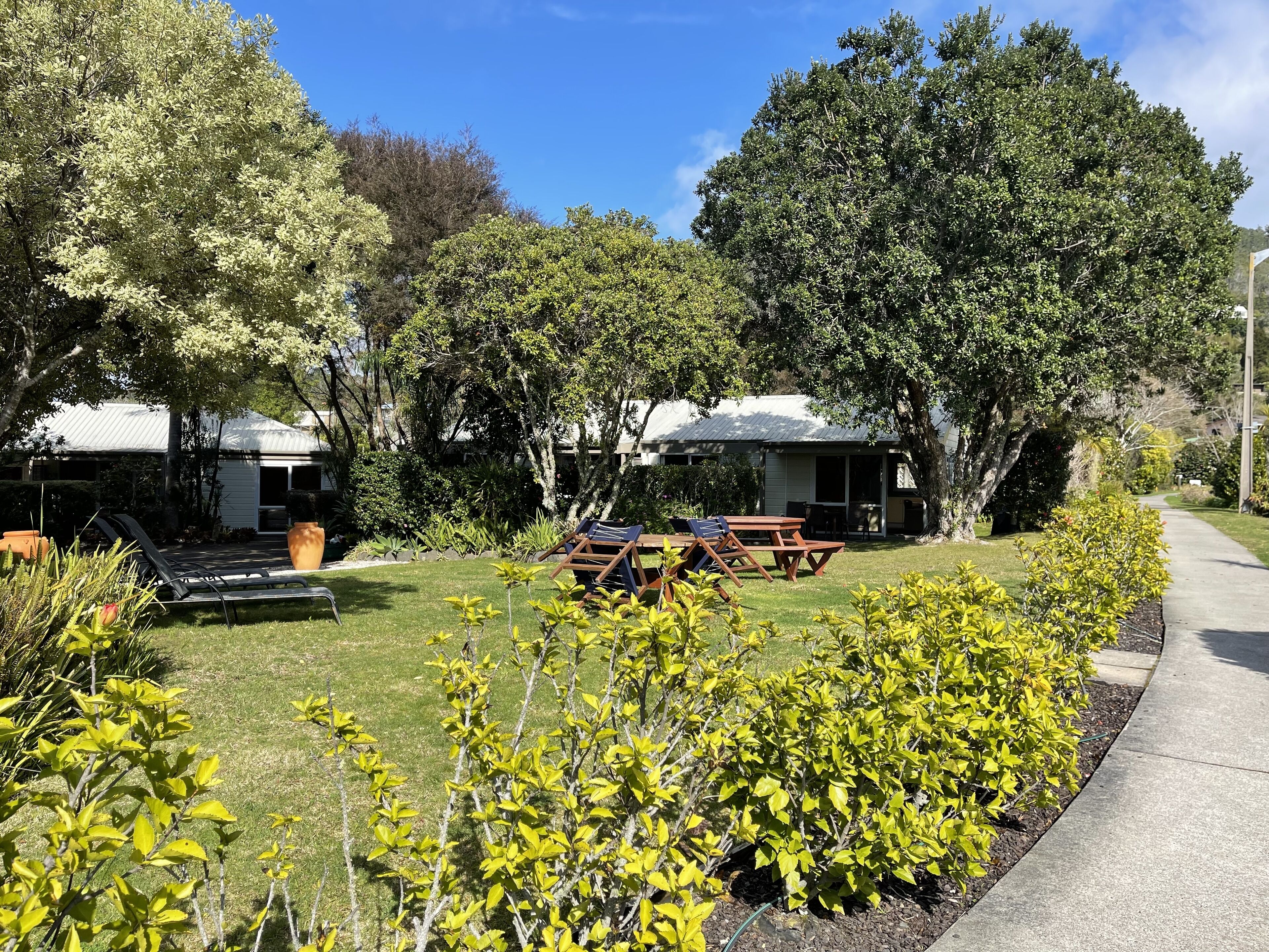 A well-maintained garden with picnic tables and lounge chairs, surrounded by mature trees and a clear sky.