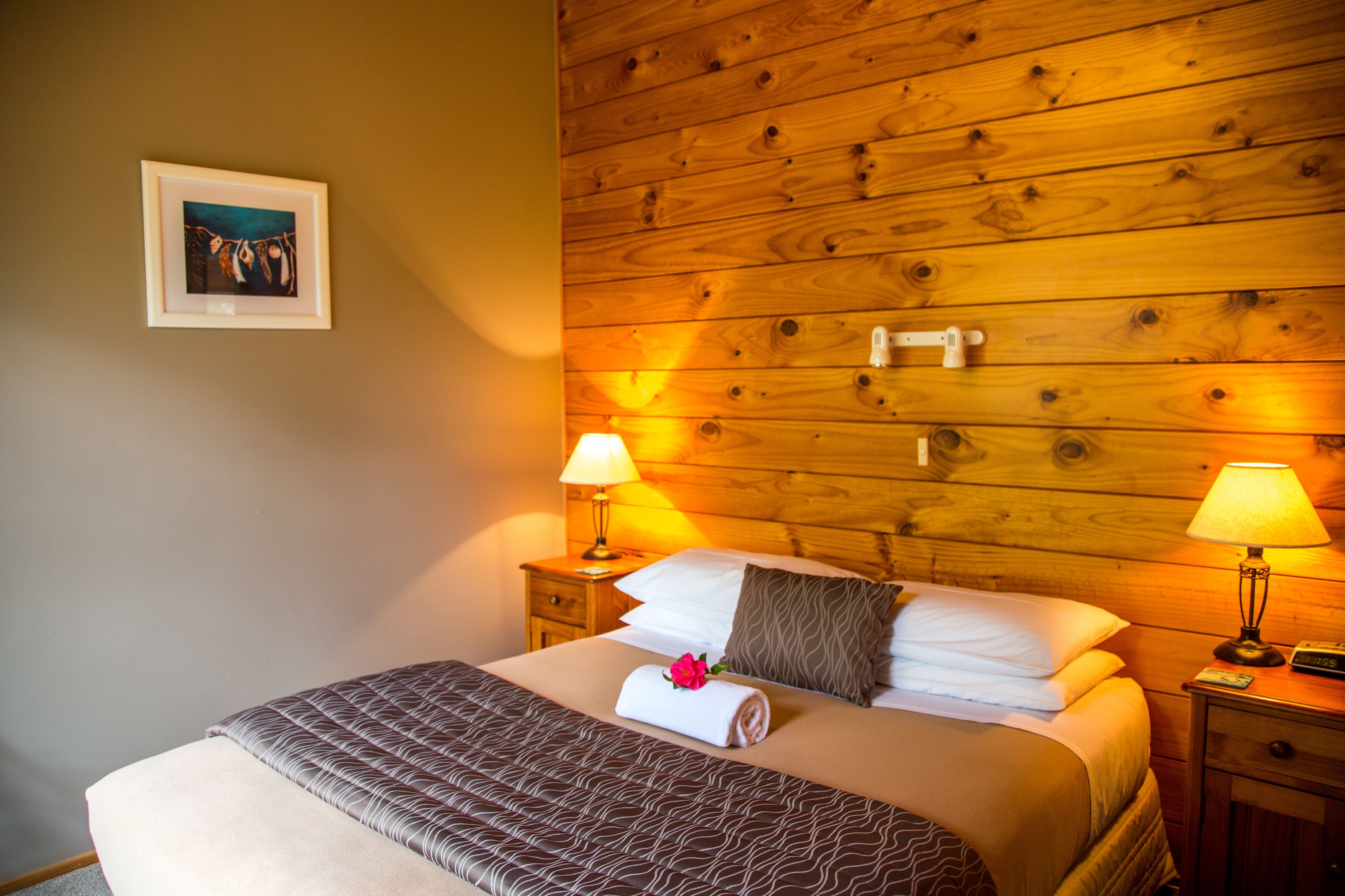 A neatly made bed with a patterned bedspread, two bedside lamps, a wooden headboard, and a framed picture on the wall.