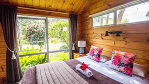 A wooden cabin bedroom with a large bed, floral pillows, a bedside lamp, and a view of greenery outside. - Tatahi Lodge Beach Resort (Hahei)
