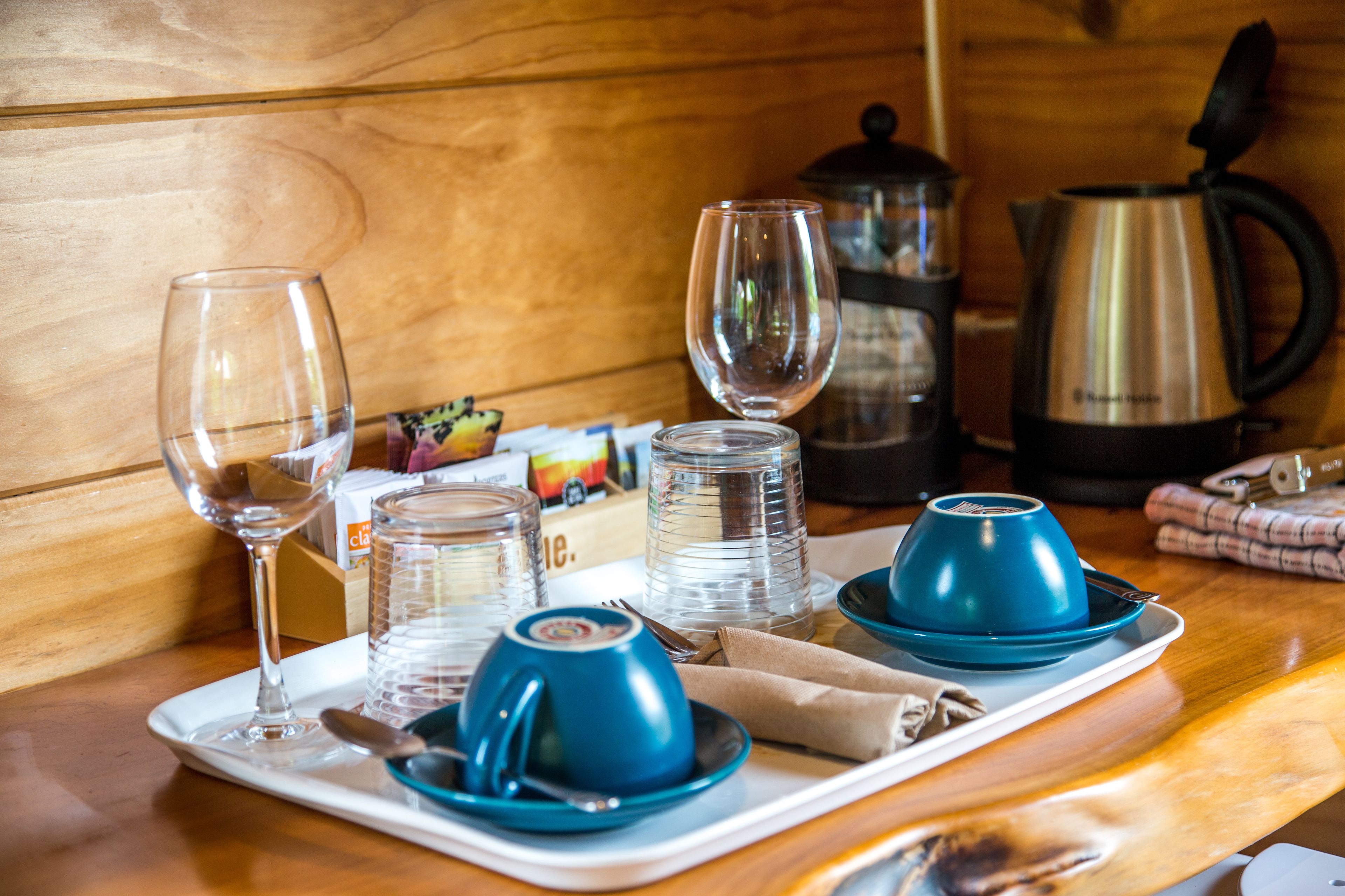 Una mesa de madera con una bandeja que contiene una copa de vino, una taza azul, un tarro de vidrio y una cafetera azul.
