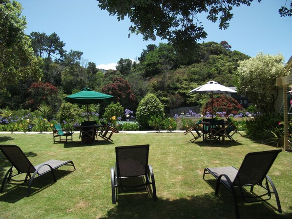 An outdoor seating area with lawn chairs and tables, surrounded by lush greenery and flowering plants. - Tatahi Lodge Beach Resort (Hahei)