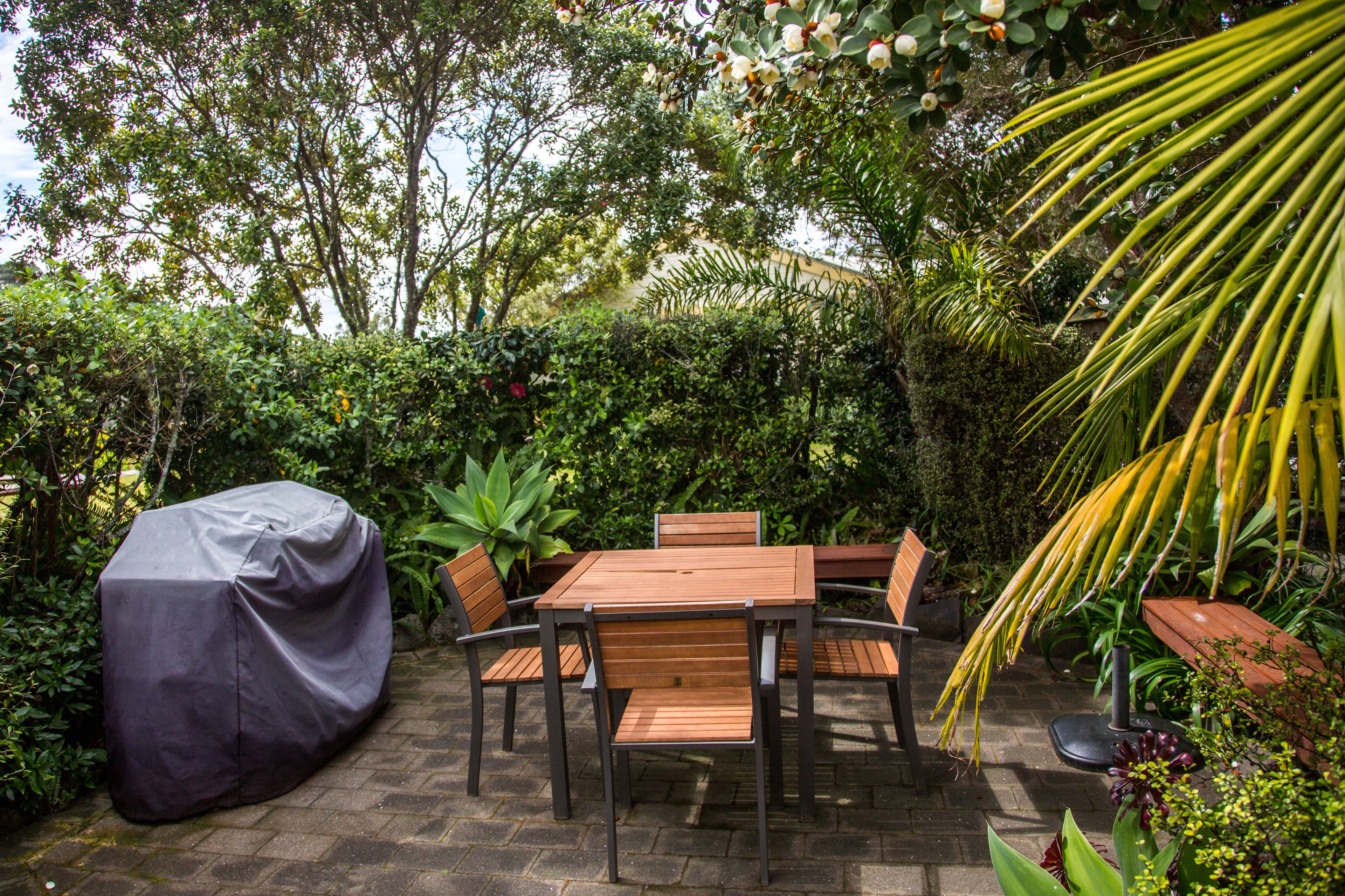 An outdoor patio with a covered table and chairs, surrounded by lush greenery and a palm tree.