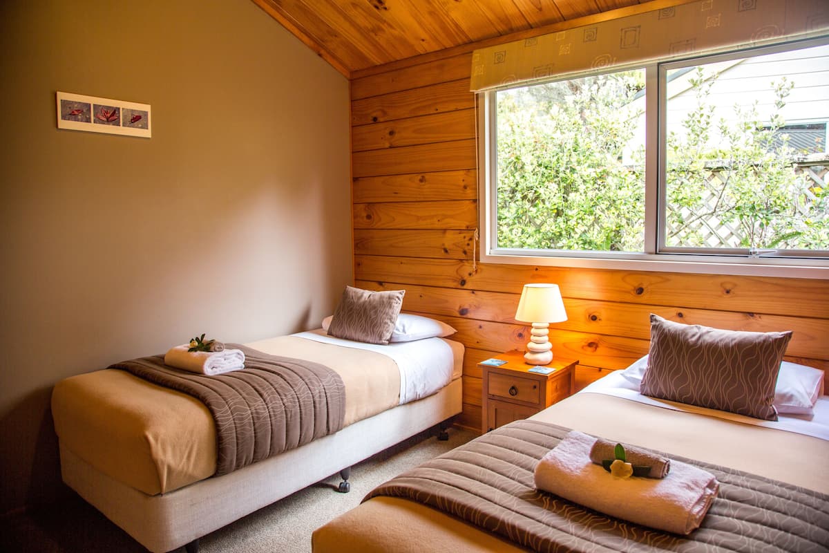 A room with two beds, wooden walls, a window with a view of greenery, and a small bedside table with a lamp.