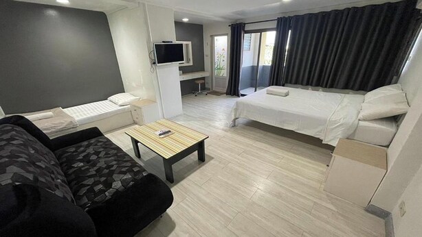 A compact hotel room with a sofa, a bed, a small table, a TV, and a balcony with curtains. - Sakun Place (Samut Prakan)