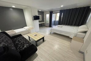 A compact hotel room with a sofa, a bed, a small table, a TV, and a balcony with curtains. - Sakun Place (Samut Prakan)
