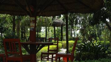 A covered outdoor seating area with a table and chairs, surrounded by lush greenery.