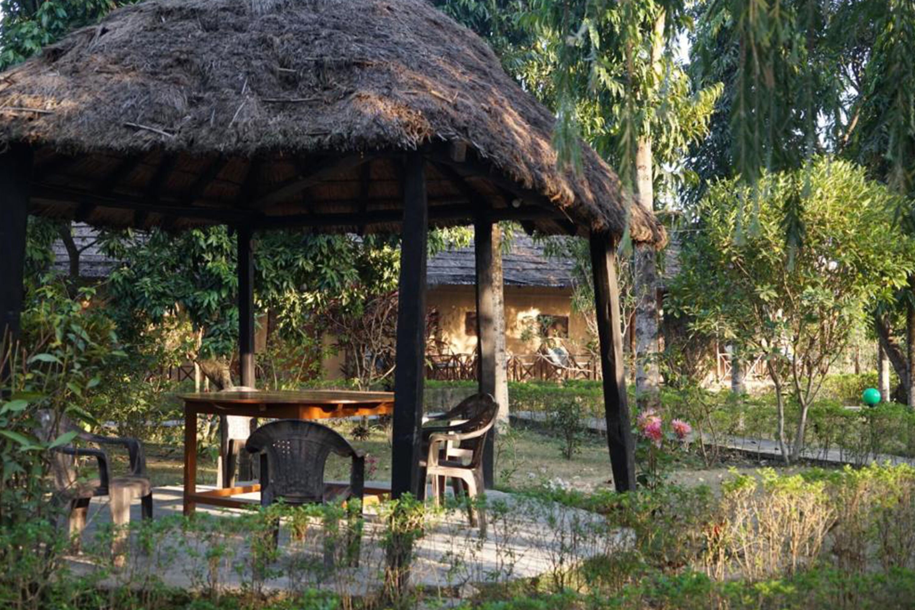 A thatched-roof gazebo with a table and chairs, surrounded by greenery and trees.