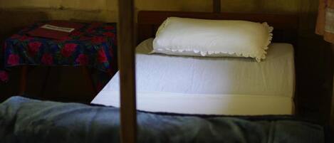 A single bed with a white mattress and pillow, a small table with a floral tablecloth, and a chair.