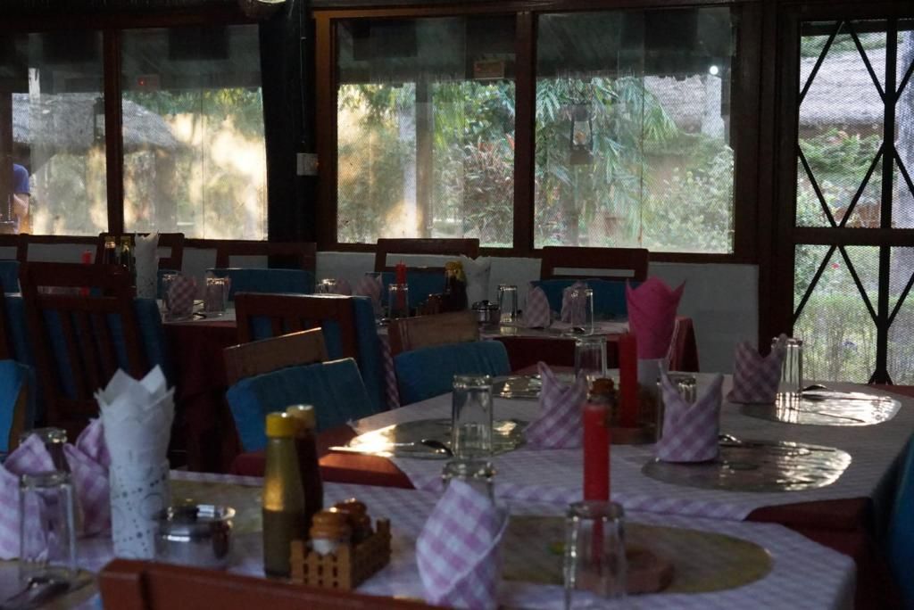 Restaurante com mesas preparadas para refeições, exibindo vista para vegetação através de grandes janelas.