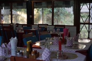 Restaurante com mesas preparadas para refeições, exibindo vista para vegetação através de grandes janelas.