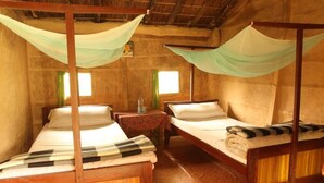 A room with two wooden beds, each with a canopy. There are two windows with curtains, a small table with a floral tablecloth, and a wooden floor. - Forest Hideaway Resort PVT LTD (Bardiya National Park)