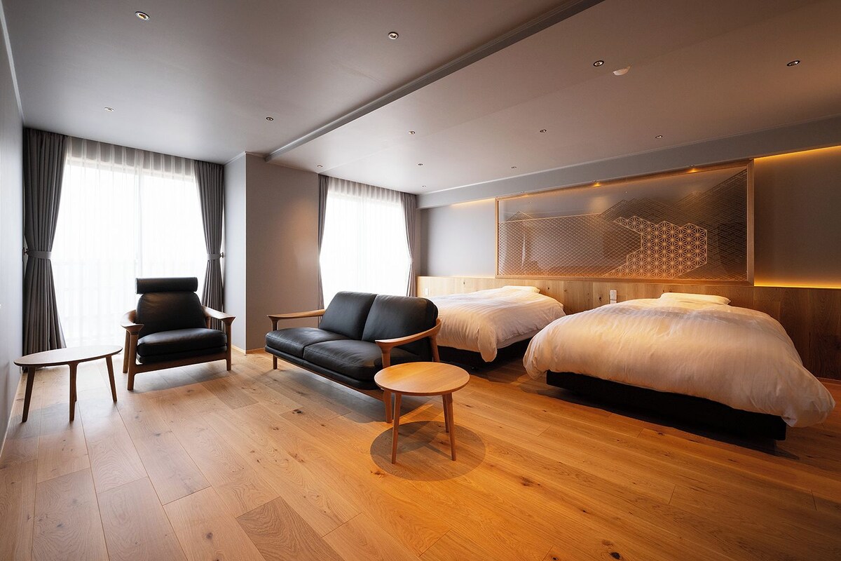 A modern hotel room with a sofa, two beds, a small table, and a decorative wall panel.