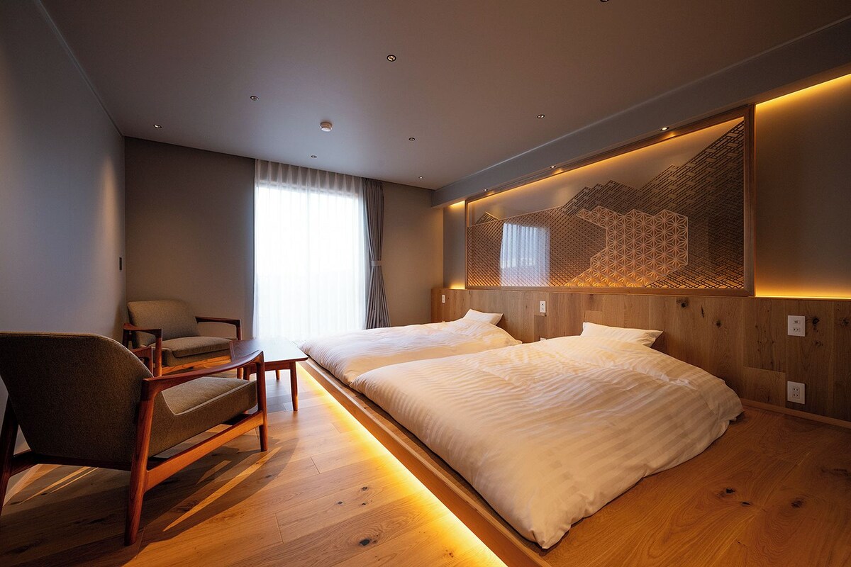 A modern hotel room with a wooden floor, a large bed, a desk with a chair, and a large window with curtains.