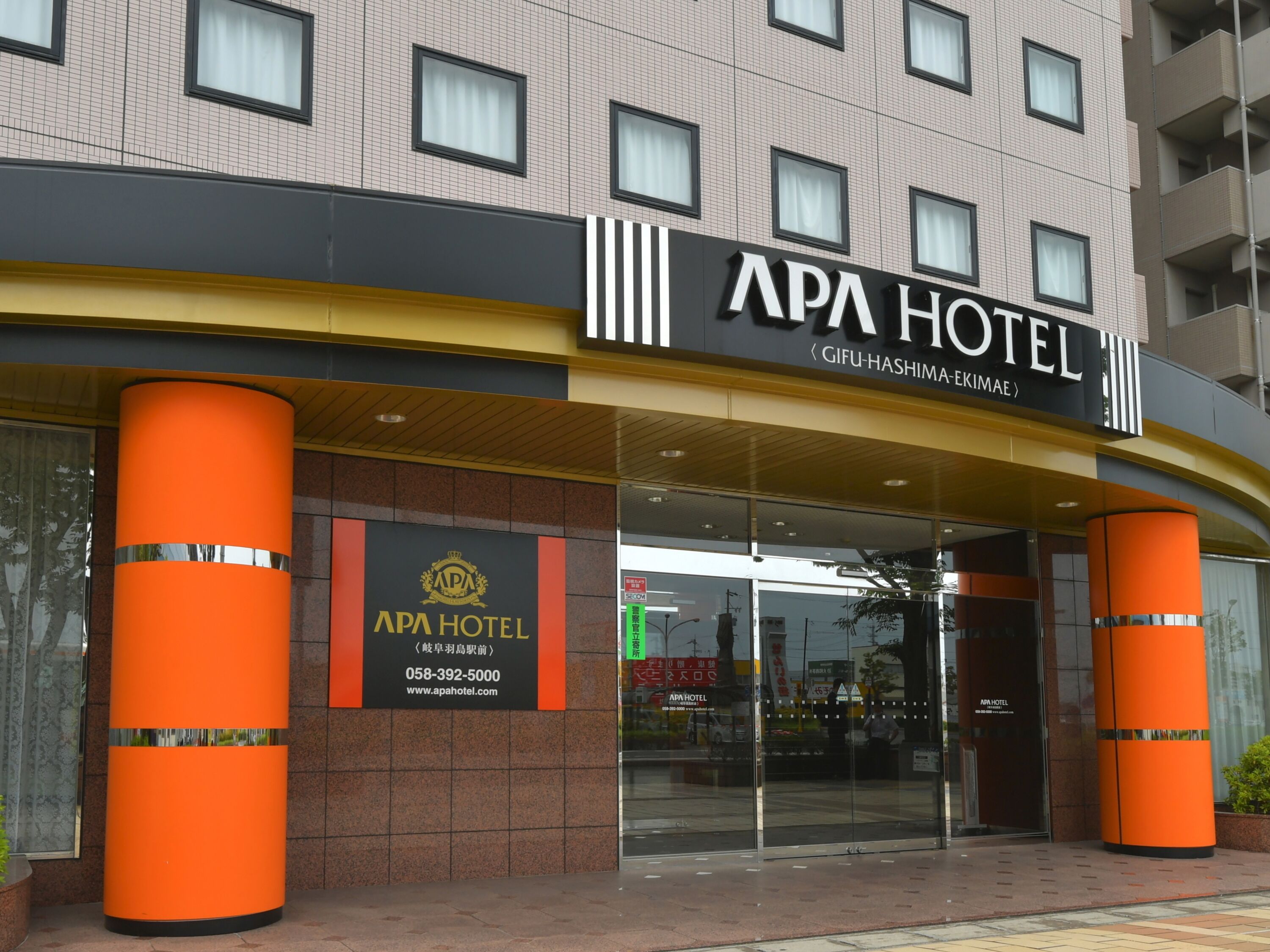 Exterior del hotel APA, con una entrada destacada, grandes pilares naranja y fachada de vidrio transparente.