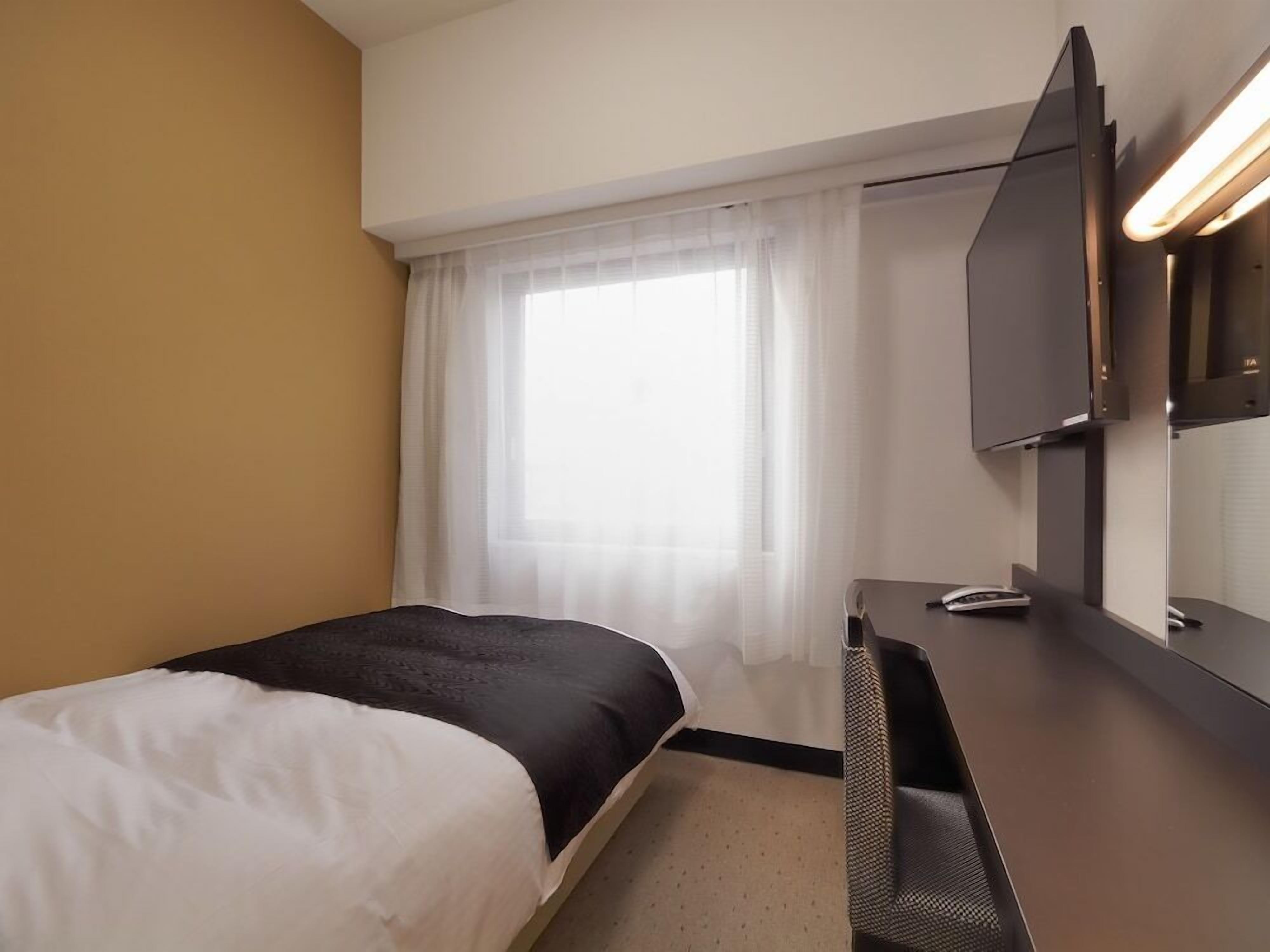 Ett hotellrum med en säng, ett skrivbord med en spegel, en tv och ett fönster med tunna, genomskinliga gardiner.