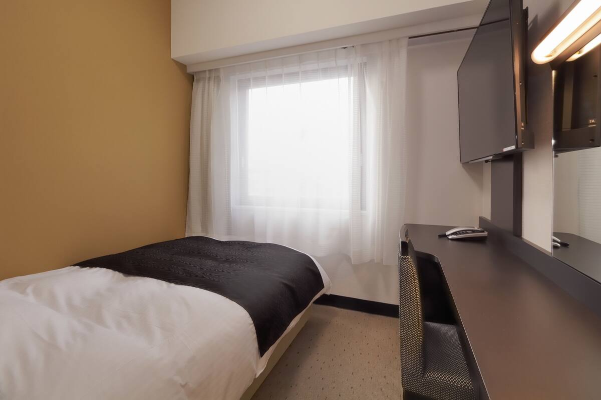 Une chambre d’hôtel avec un lit, un bureau doté d’un miroir, une télévision et une fenêtre avec des rideaux légers.
