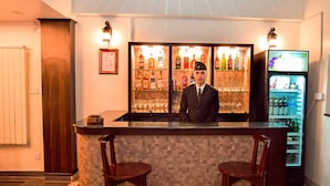 Una zona de bar con un cantinero, una barra azulejada, dos taburetes de madera, una estantería con botellas y un refrigerador.
