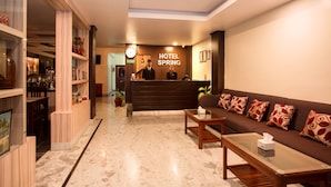 Un vestíbulo de hotel con mostrador de recepción, un sofá con cojines estampados, una mesa de centro y una estantería con libros y objetos decorativos.