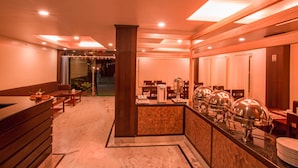 Vestíbulo de hotel con mostrador de recepción, zona de estar, bufé y equipo para servicio de alimentos, y un ventanal con cortinas.