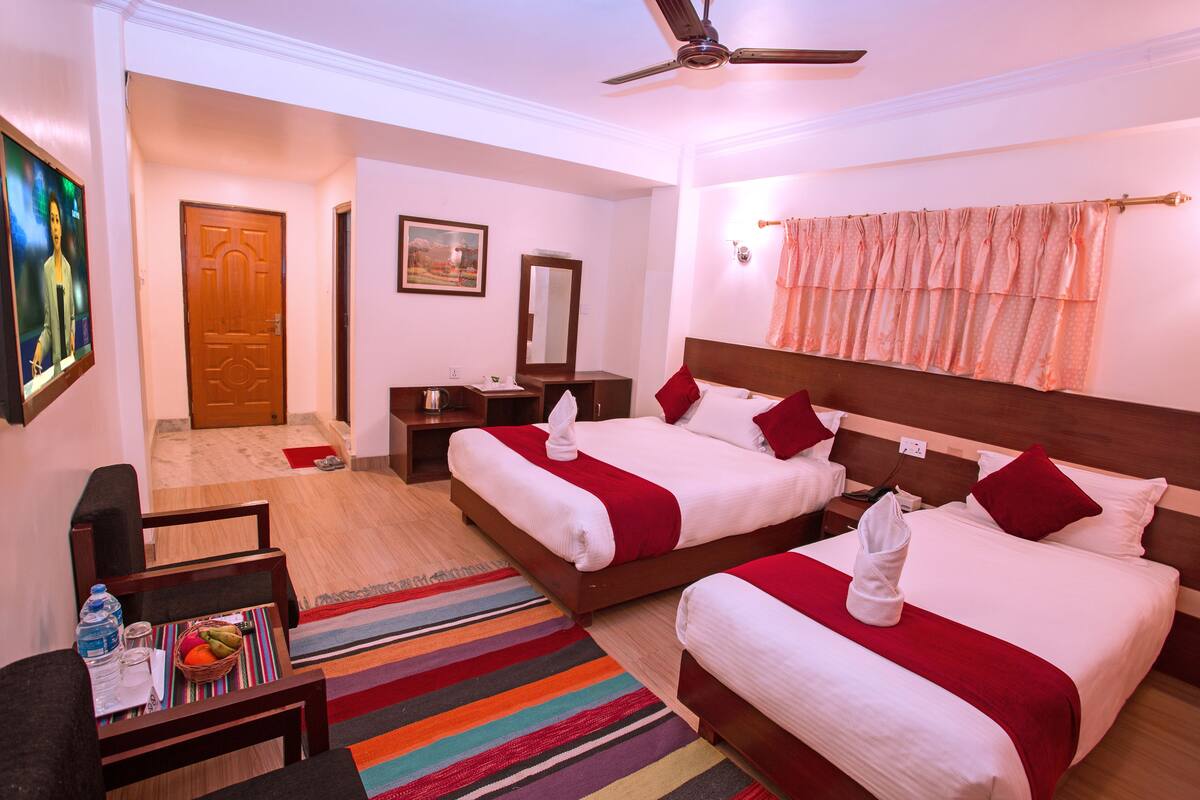 Habitación de hotel con dos camas, ventilador de techo, cabecero de madera, alfombra a rayas de colores, una pequeña mesa con frutero y un cuadro en la pared.