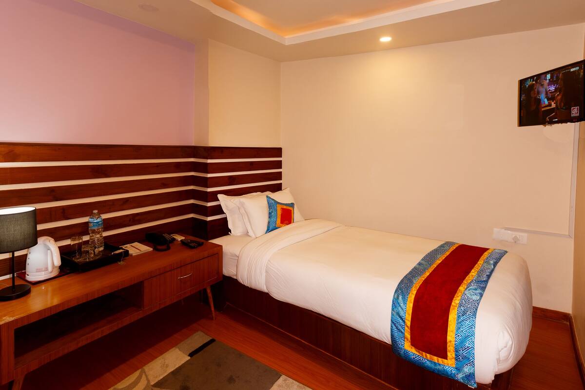 Una habitación de hotel con cabecera de madera, cama con ropa blanca y un plaid colorido, mesita de noche con lámpara, control remoto y una botella de agua.