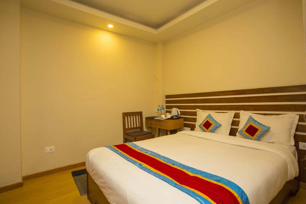 Habitación de hotel con cama, mesita de noche, una silla y una pequeña mesa con una botella de agua y una taza.