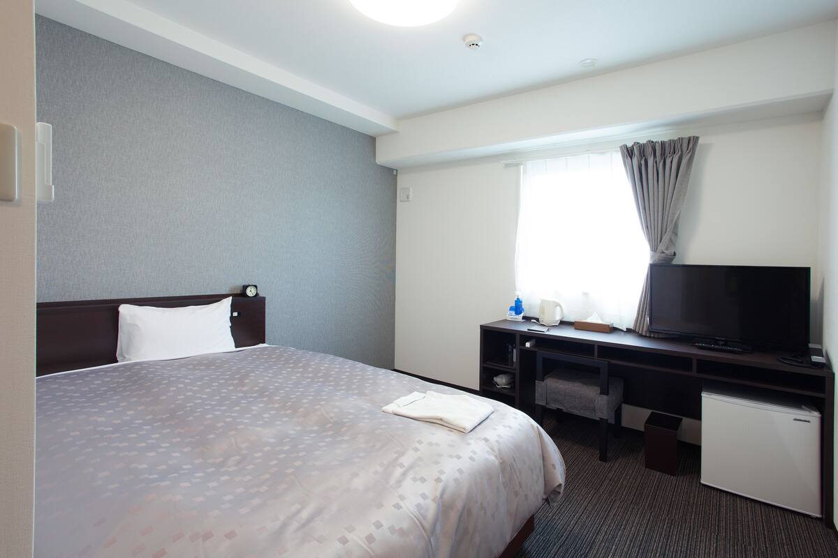 Ein Hotelzimmer mit Bett, Schreibtisch, Fernseher, einer Minikühlschrank und einem Vorhangfenster.